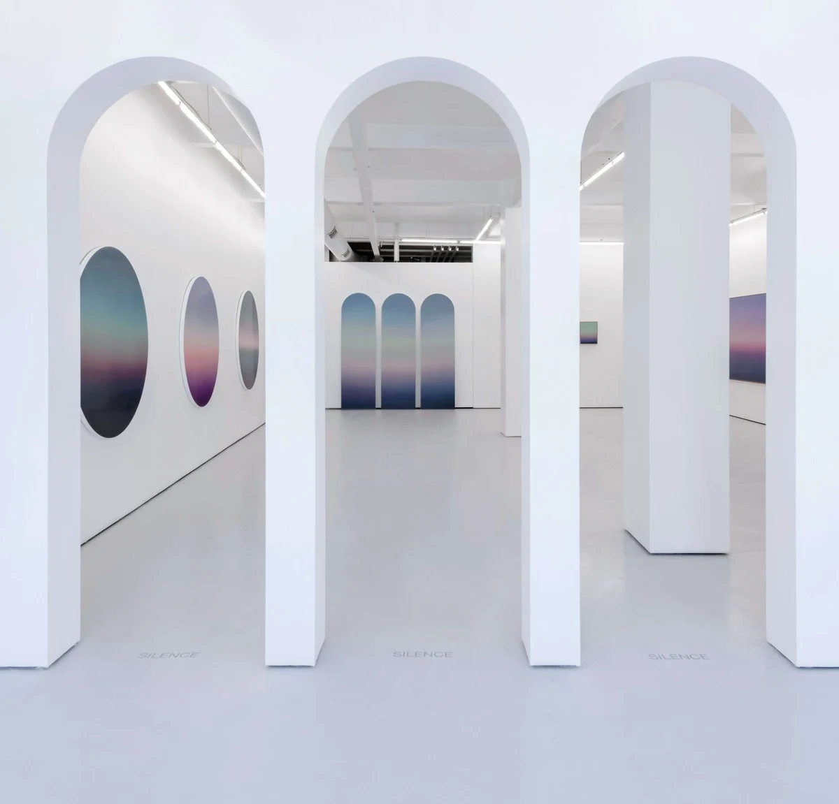 A white gallery of tall arched openings, a circular painting centred on the wall beyond each arch.