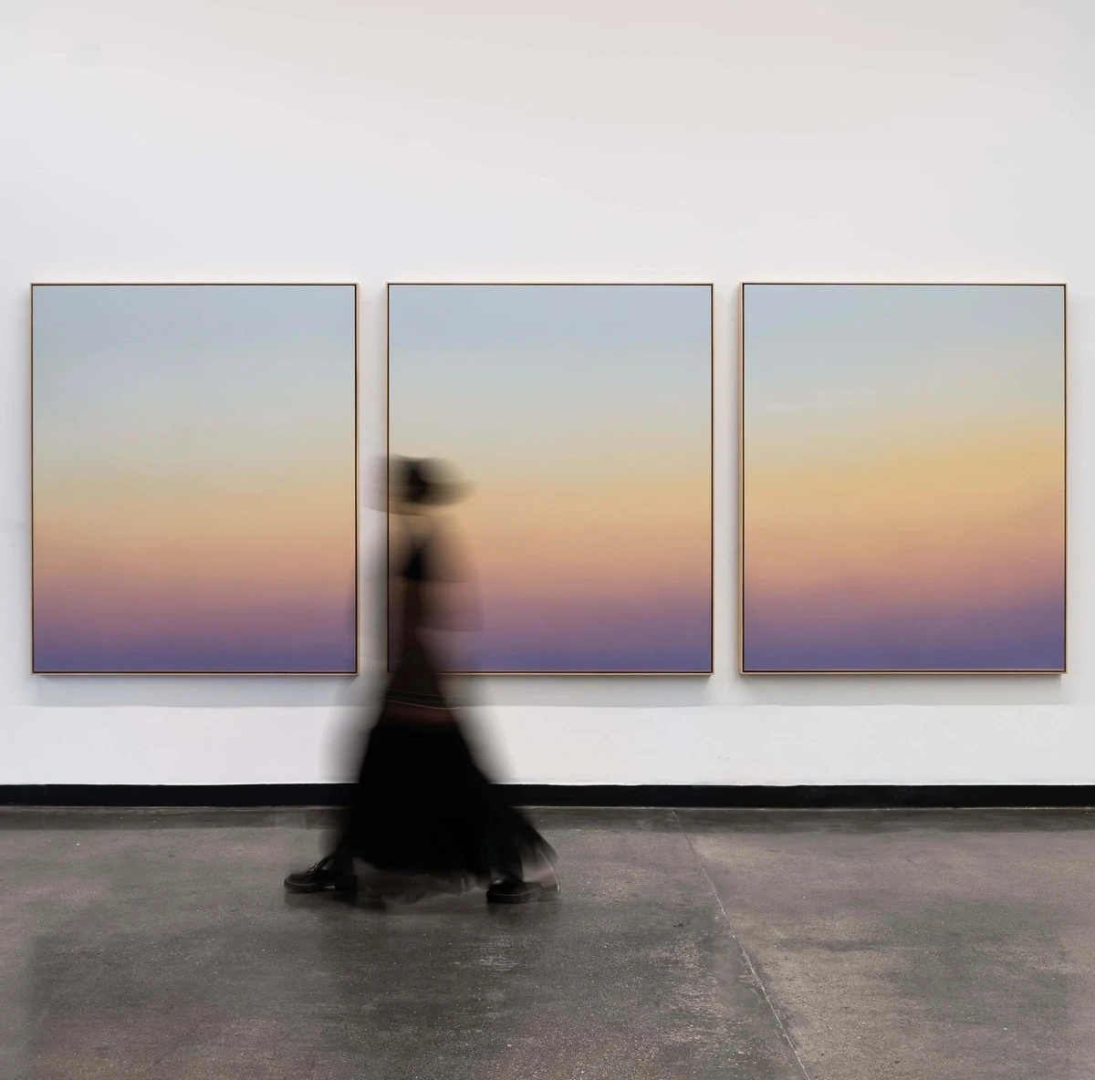 Three wide canvases of graded amber and violet along a concrete-floored wall; a figure in black walks past, blurred.