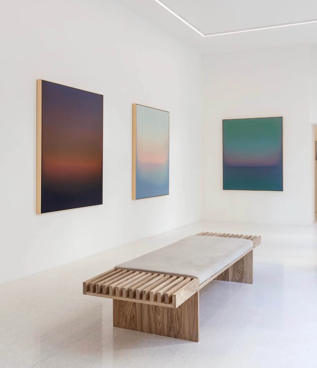 A pale gallery hung with three paintings, a slatted wooden bench along the wall beneath them.