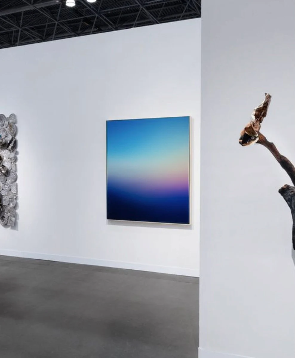 An art fair booth, a blue and rose painting on the end wall between a pale relief and a dark sculpture.