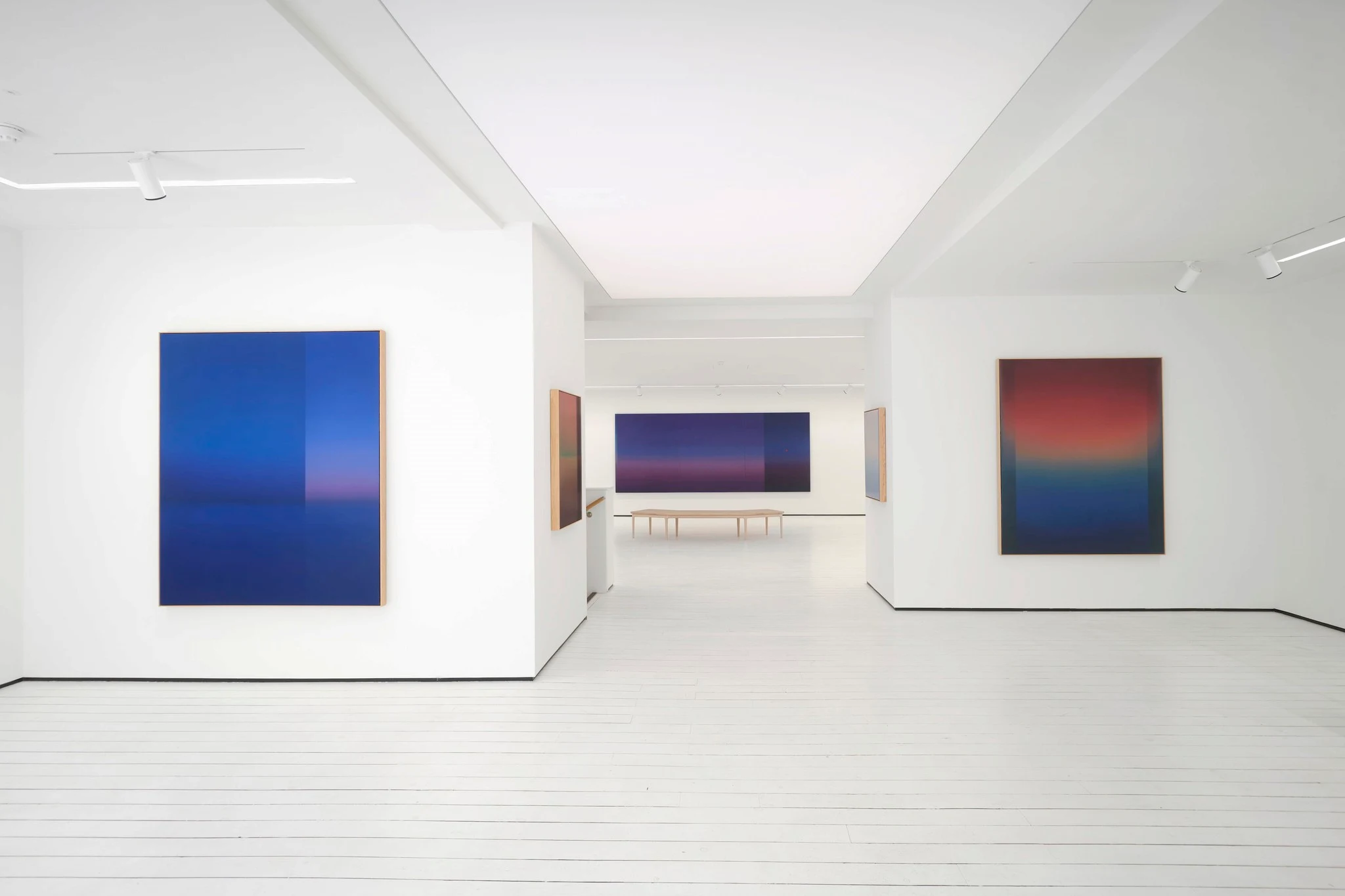 A white gallery with a deep blue painting on the near wall, the room opening past it to further work.