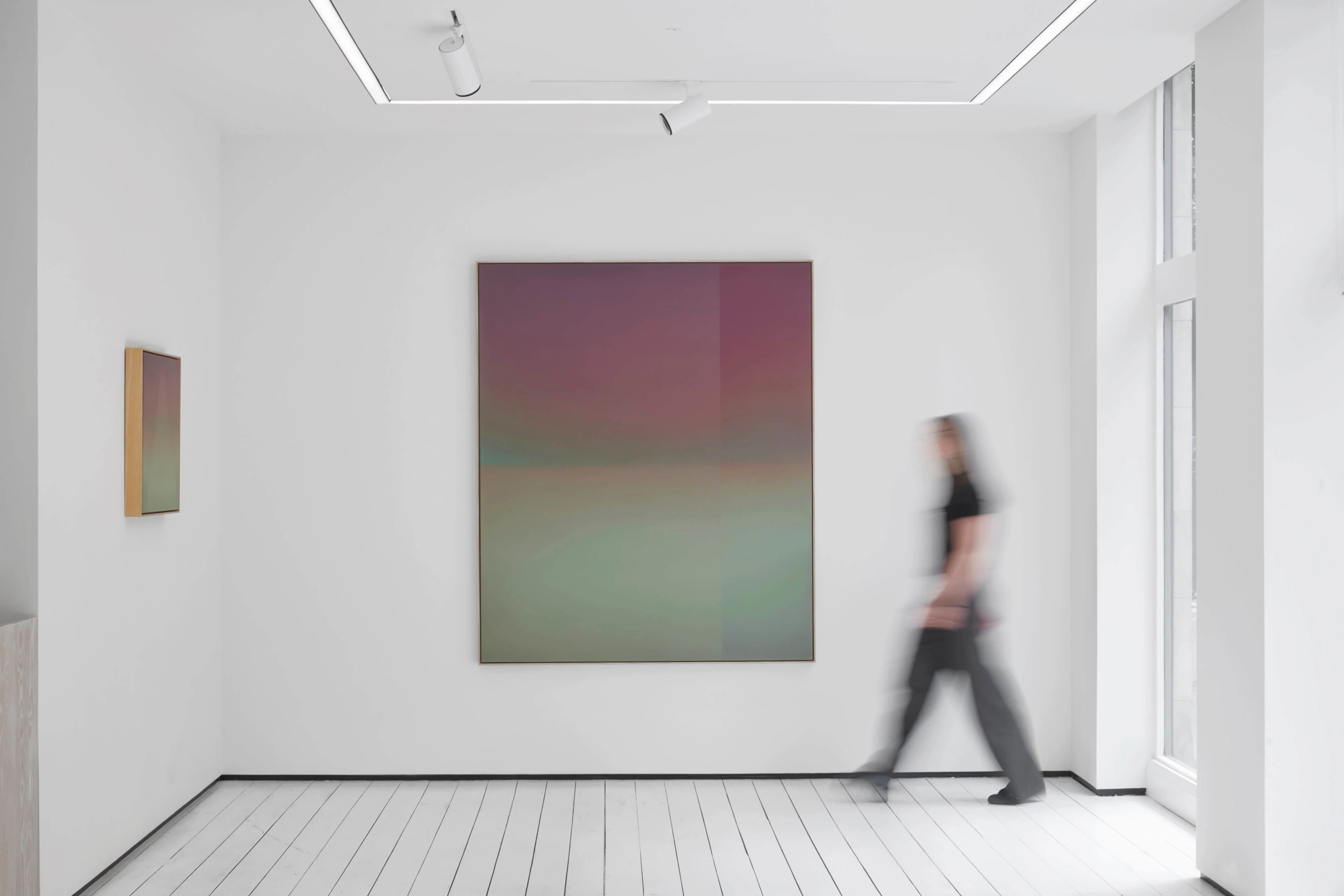 A tall painting in a white gallery, dusky rose above melting into pale green along a soft horizon; a visitor walks past, blurred.