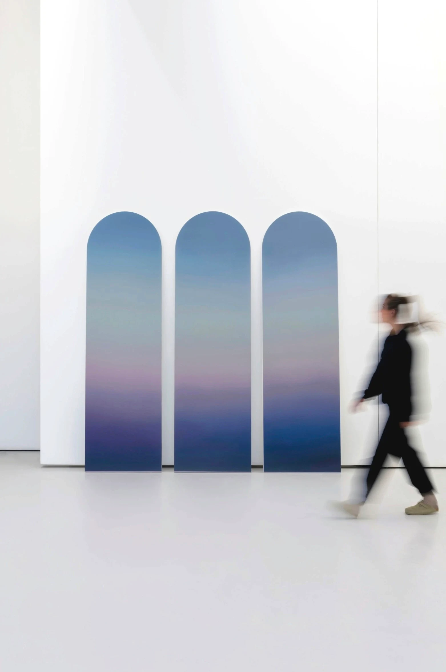 Three arch-topped panels leaning together against a white wall, each graded from blue through violet into deeper blue; a figure walks past.