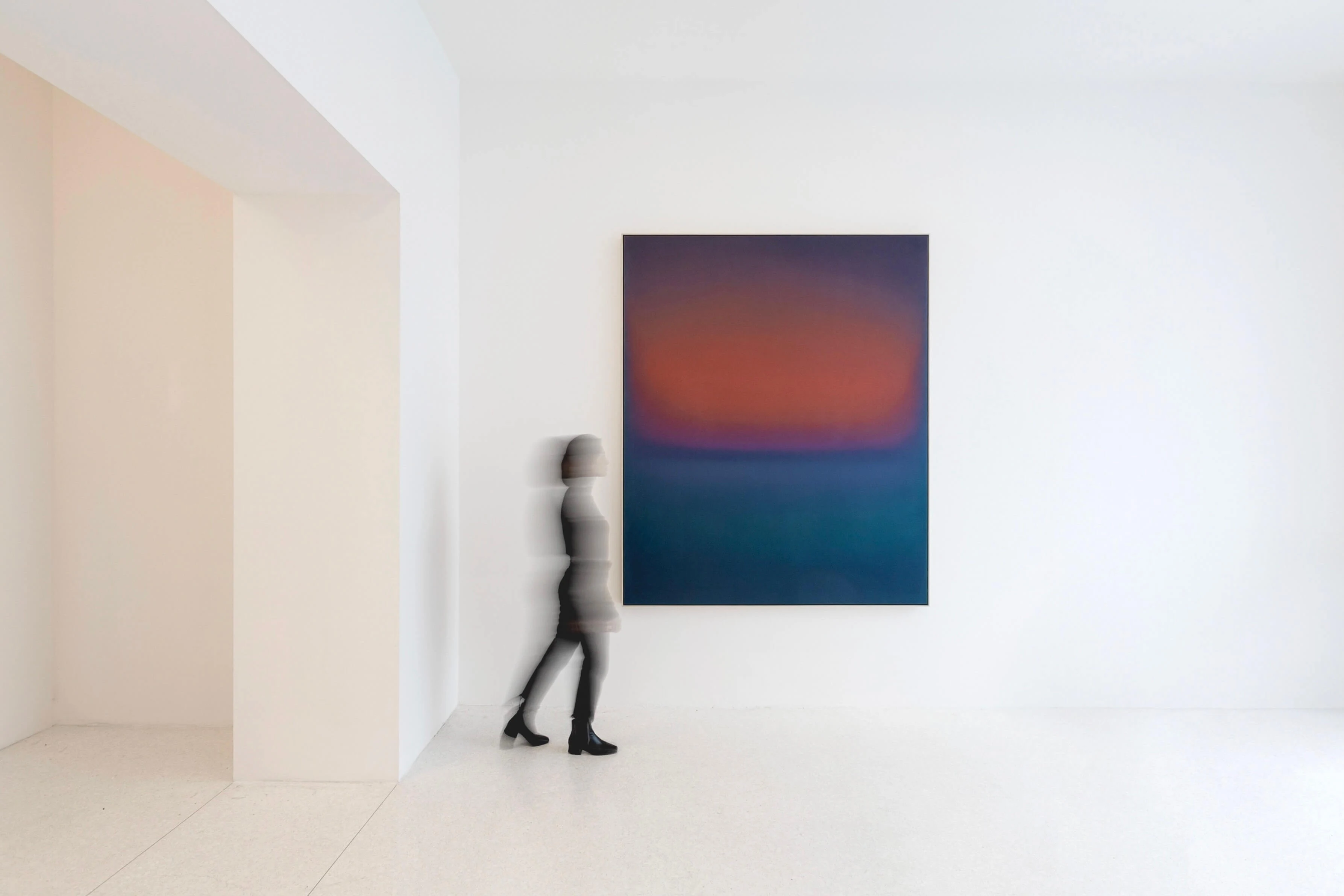 A tall canvas glowing burnt orange above deep teal, hung in a bare white room as a figure passes in front of it.