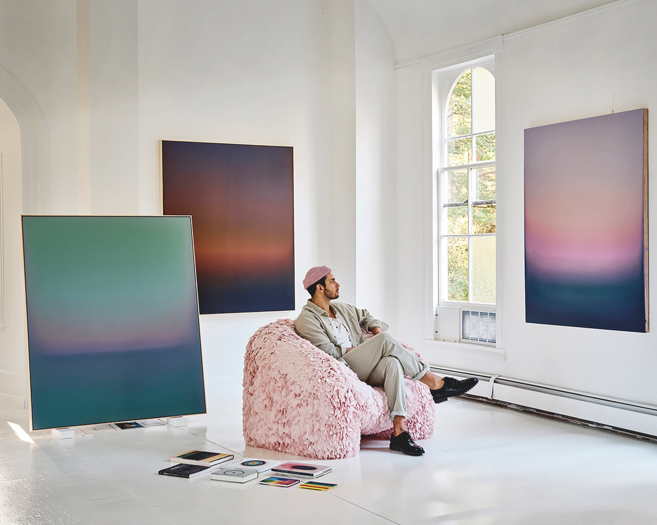 Theo Pinto seated on a pale pink chair in his studio, finished paintings leaning and hung around him beside a tall arched window.