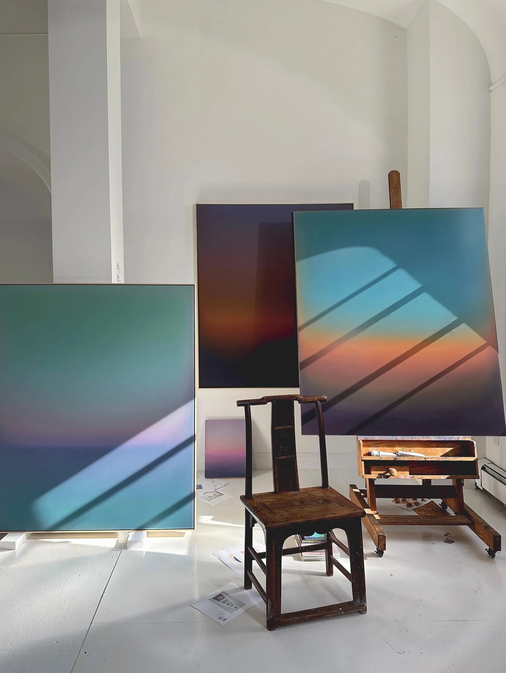 Canvases leaning against a studio wall beside a wooden chair, one teal and one graded through rose into blue.