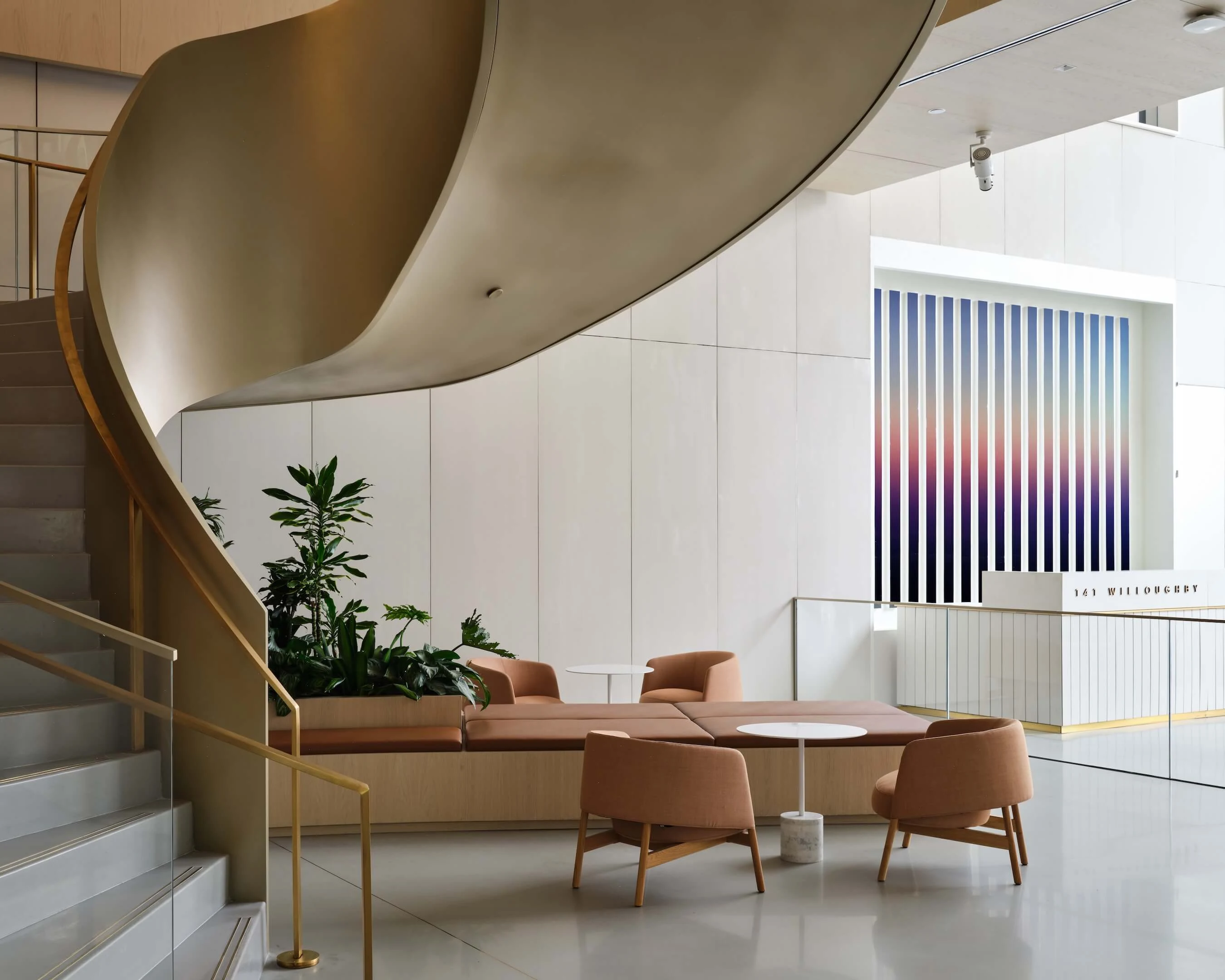 A tall striped work of graded blues and warm pinks installed in a light-filled lobby beside a curving brass staircase.